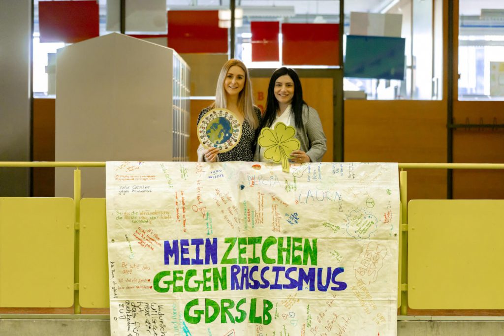 Die zwei Ethik-Lehrerinnen stehen vor einem Plakat mit dem Titel "Mein Zeichen gegen Rassismus an der GDRSLB"
